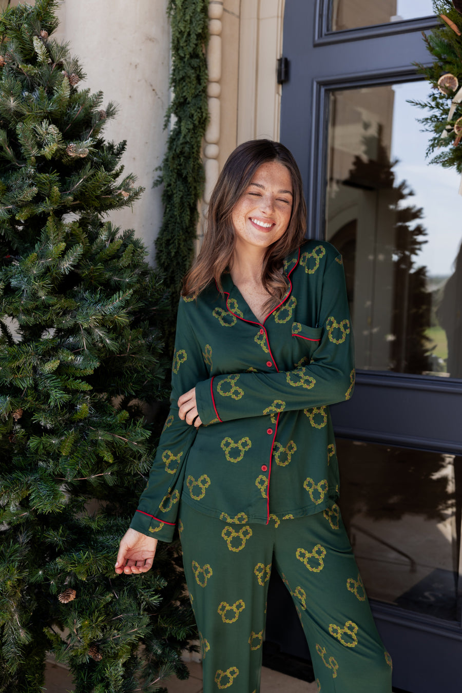 Woman wearing Long-Sleeved Women's Pajama Set in Mickey Christmas Wreath print smiling outdoors beside a Christmas tree