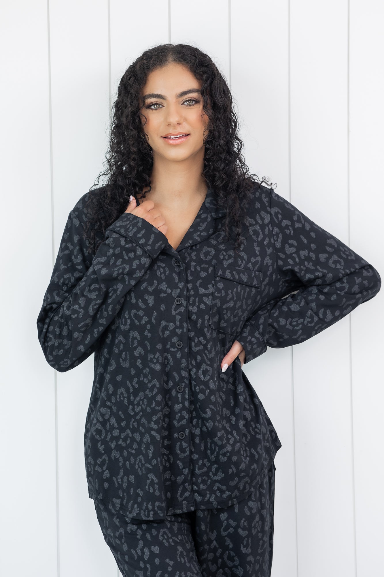 Woman wearing black button up pajamas in a monochromatic leopard print with white wooden wall backdrop