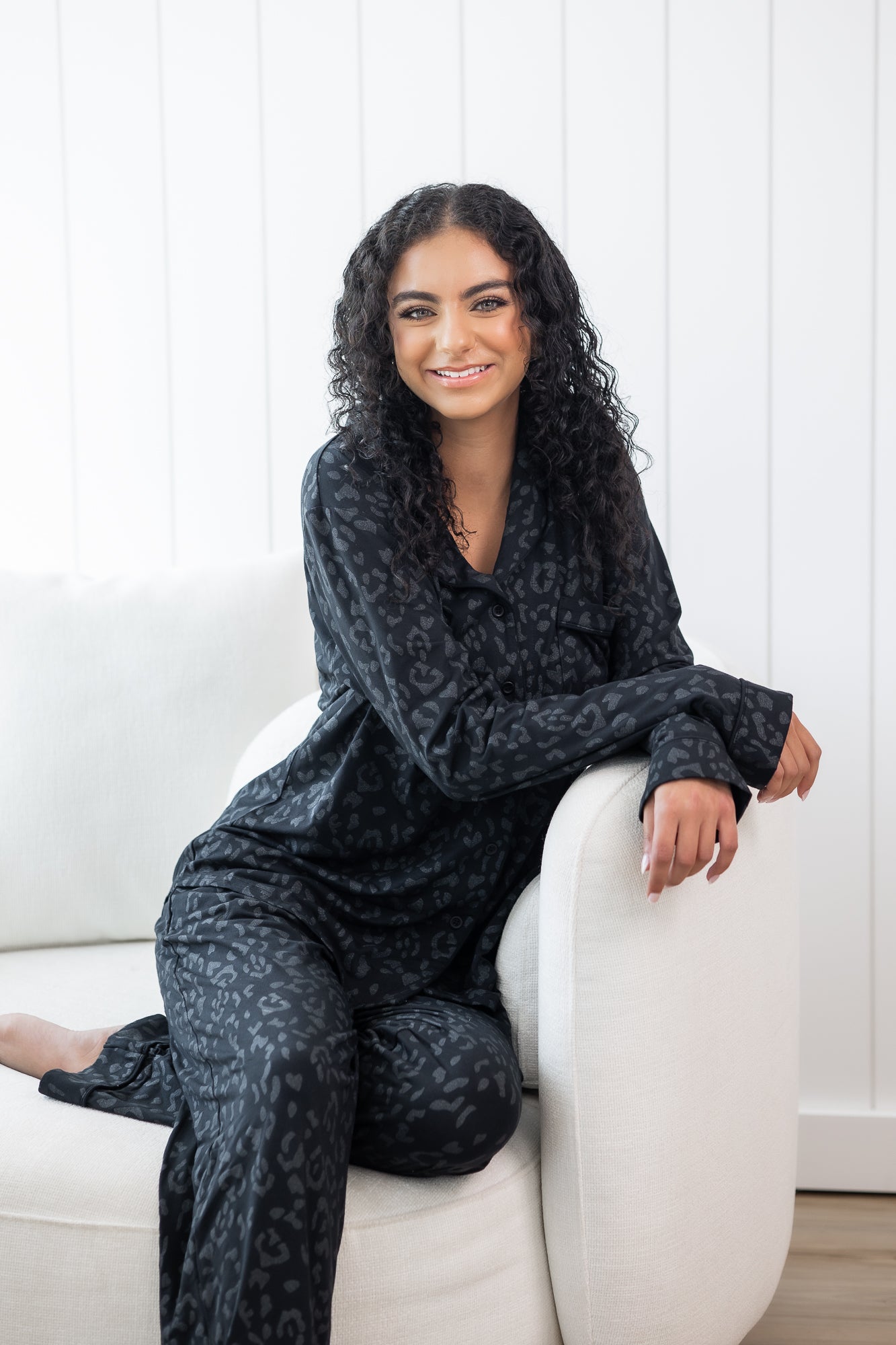Woman sitting in cream chair wearing black button up pajamas in a monochromatic leopard print