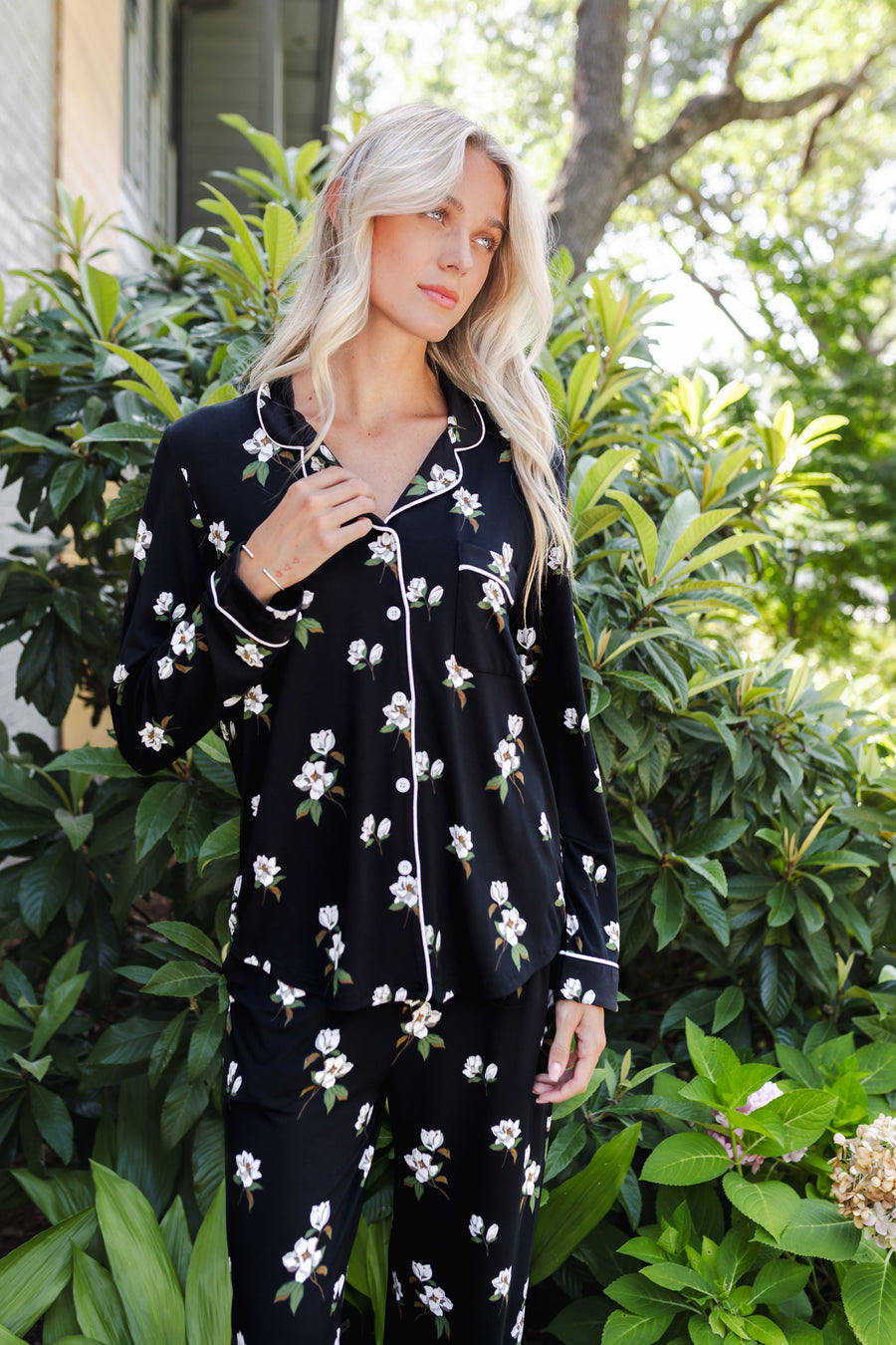 A woman wearing a black long-sleeve pajama set with a white floral print, standing outdoors in front of green foliage.