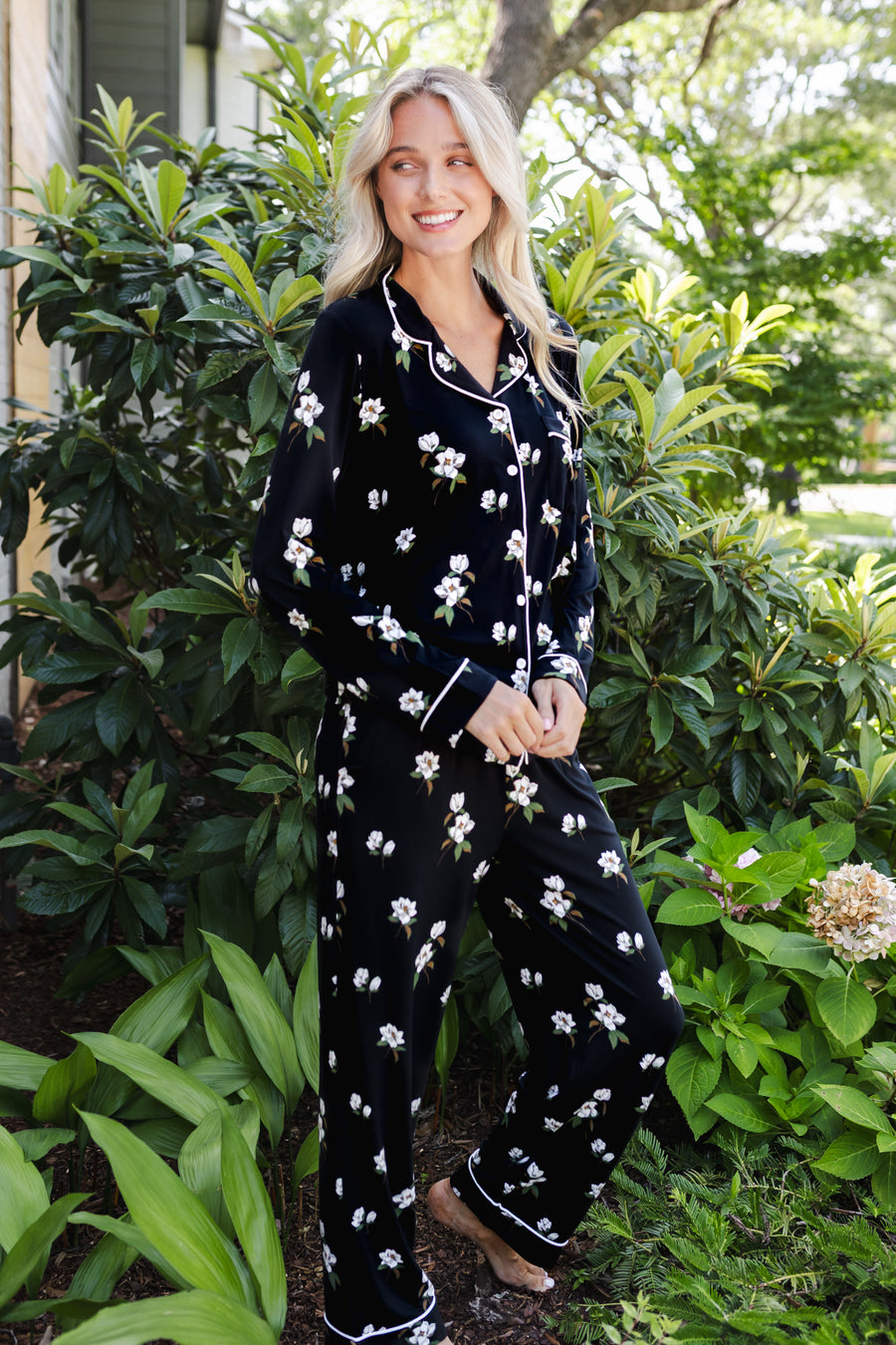 A woman wearing a black long-sleeve pajama set with a white floral print and white piping trim, standing outdoors in front of lush green garden foliage.