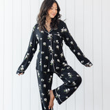Woman modeling black long sleeve button up pajamas in a magnolia print against a white wooden wall