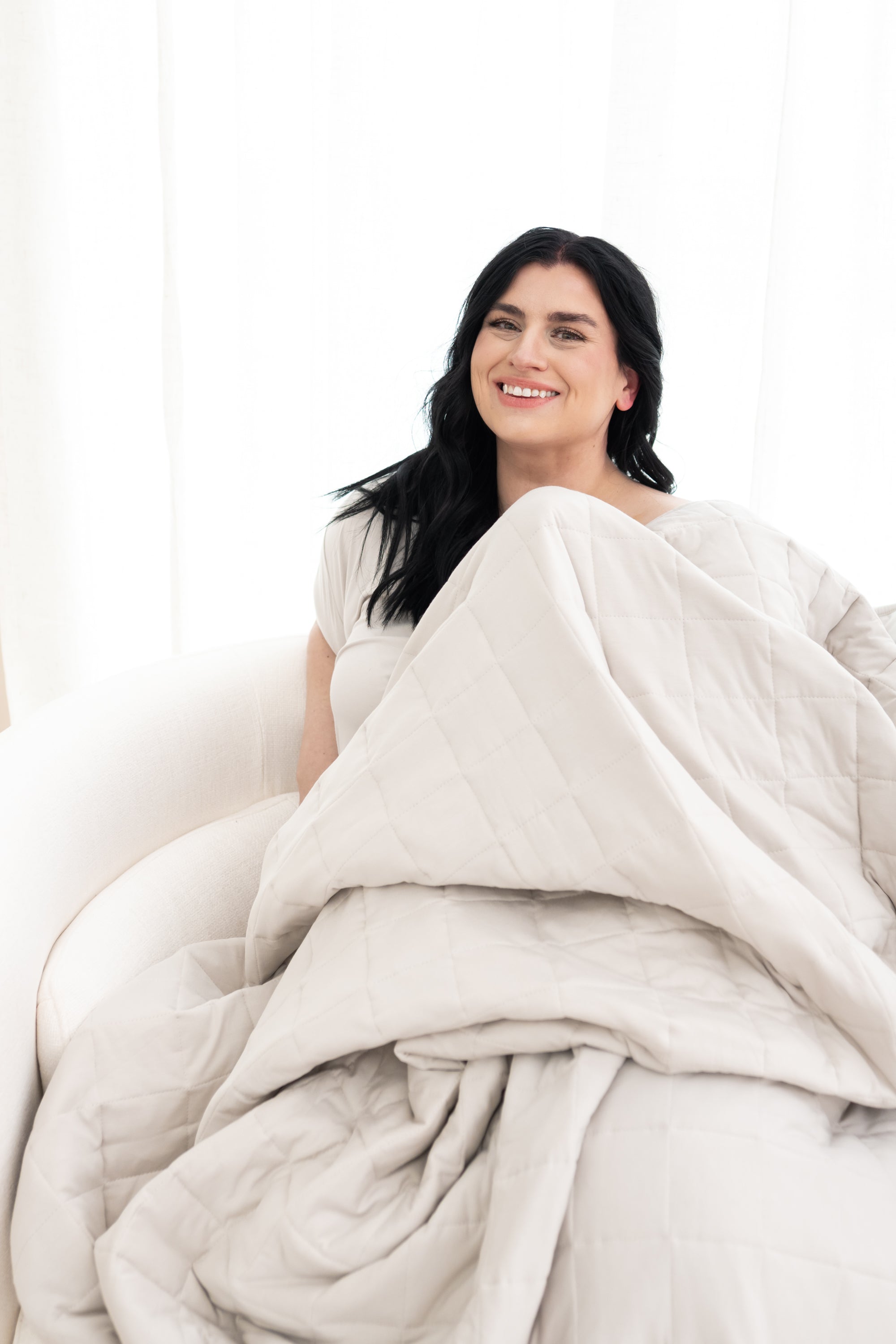 Model sitting on a chair with the Adult Quilted Blanket in Oat 2.5 on top of her