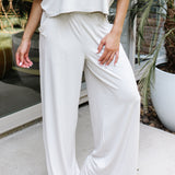 Female model standing on a sidewalk in front of a glass door of a white brick house wearing the Women's Wide Leg Pant in Oat