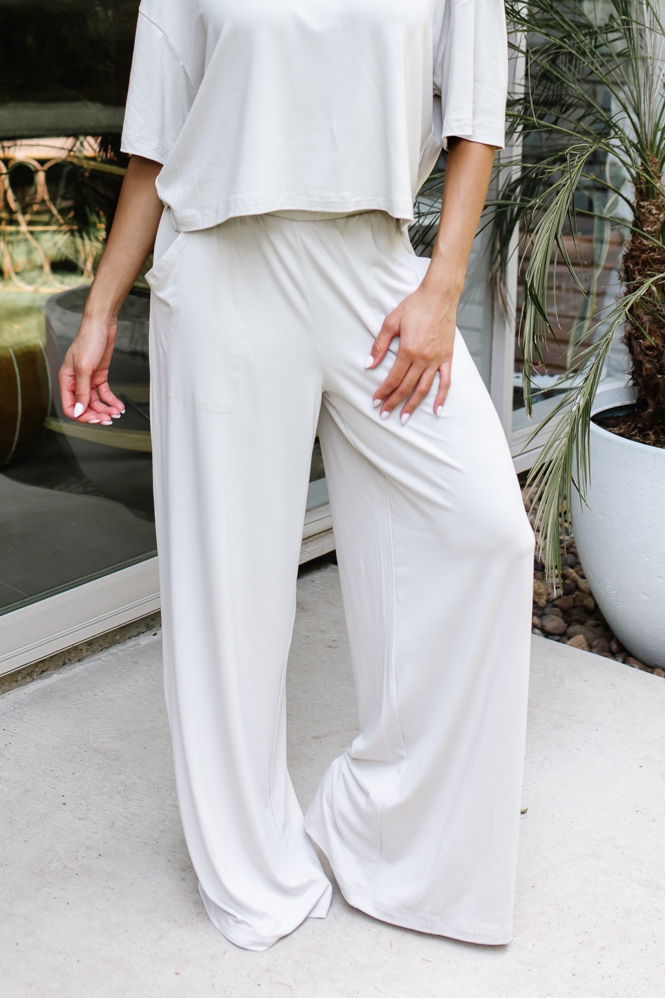 Female model standing on a sidewalk in front of a glass door of a white brick house wearing the Women's Wide Leg Pant in Oat