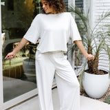 Smiling female standing on a concrete patio in front of glass windows on a white brick house wearing the Women's Wide Leg Pant in Oat and matching women's short sleeve tee