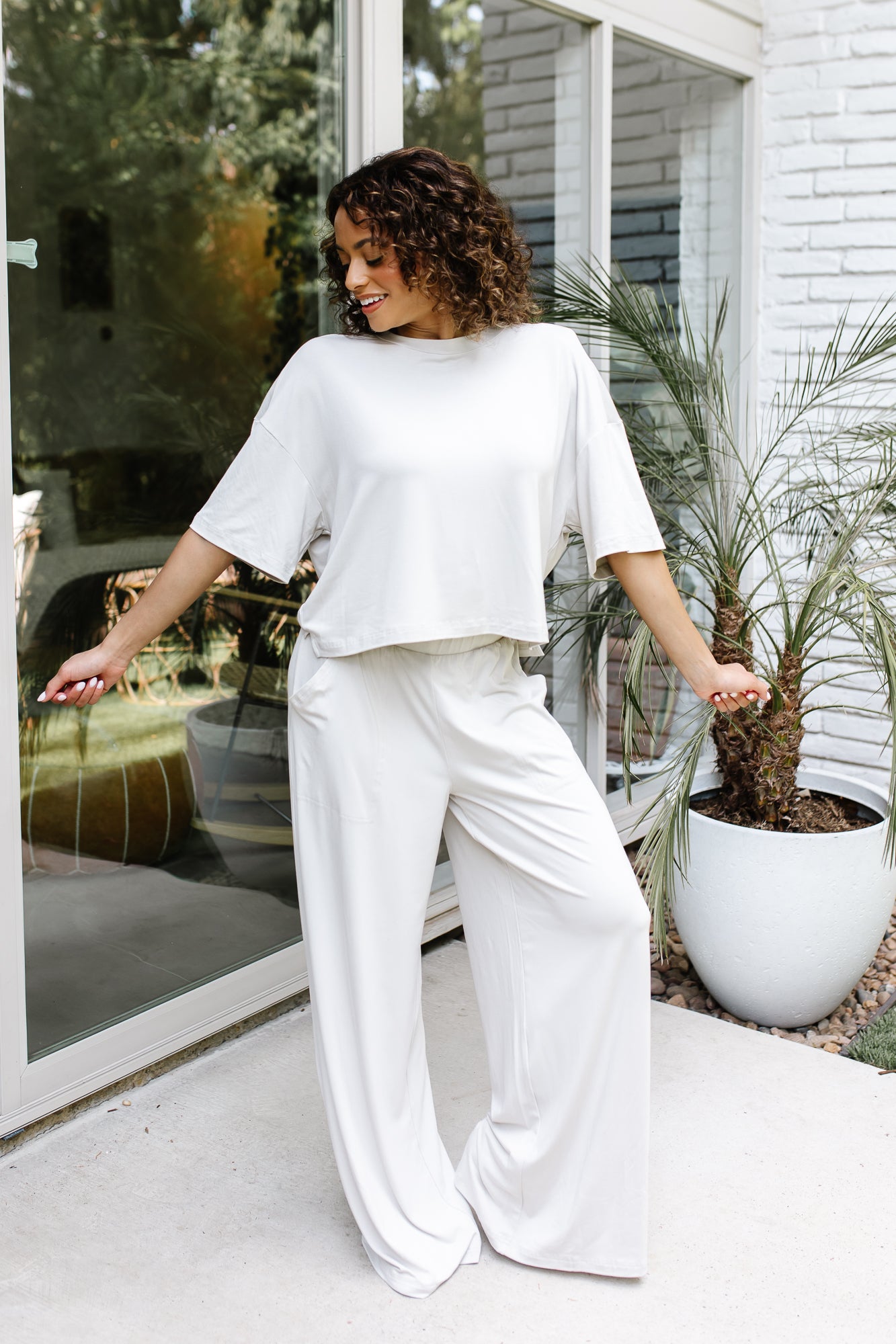 Smiling female standing on a concrete patio in front of glass windows on a white brick house wearing the Women's Wide Leg Pant in Oat and matching women's short sleeve tee