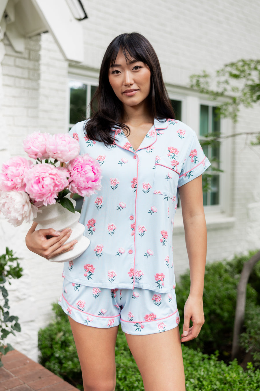 Woman outside soft smiling holding a vase of flowers in short sleeve pajamas and shorts in Peony on Breeze print.