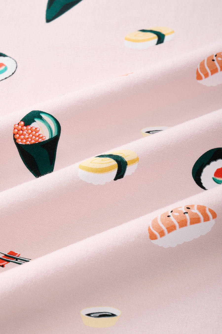 A close-up of a pink fabric with a repeating sushi print, including nigiri, maki, and sashimi motifs in yellow, green, orange, and white.