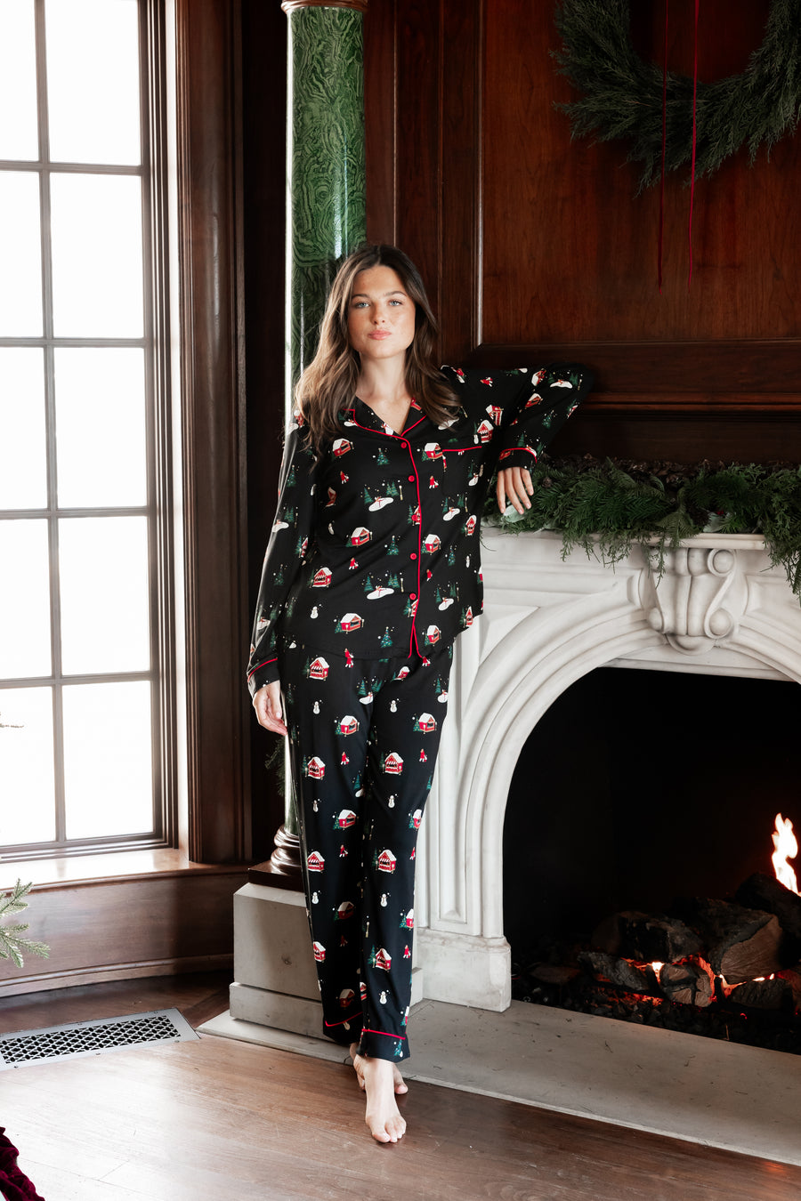 A woman wearing a black long-sleeve pajama set with a Santa Claus and snowman print, standing barefoot in front of a fireplace with a window to her left.