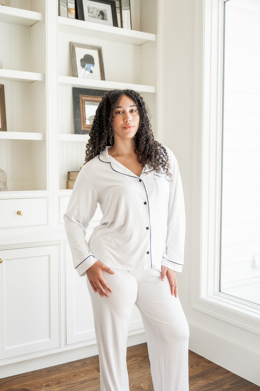 Woman wearing a white pajama set in a room with a bookshelf and window.