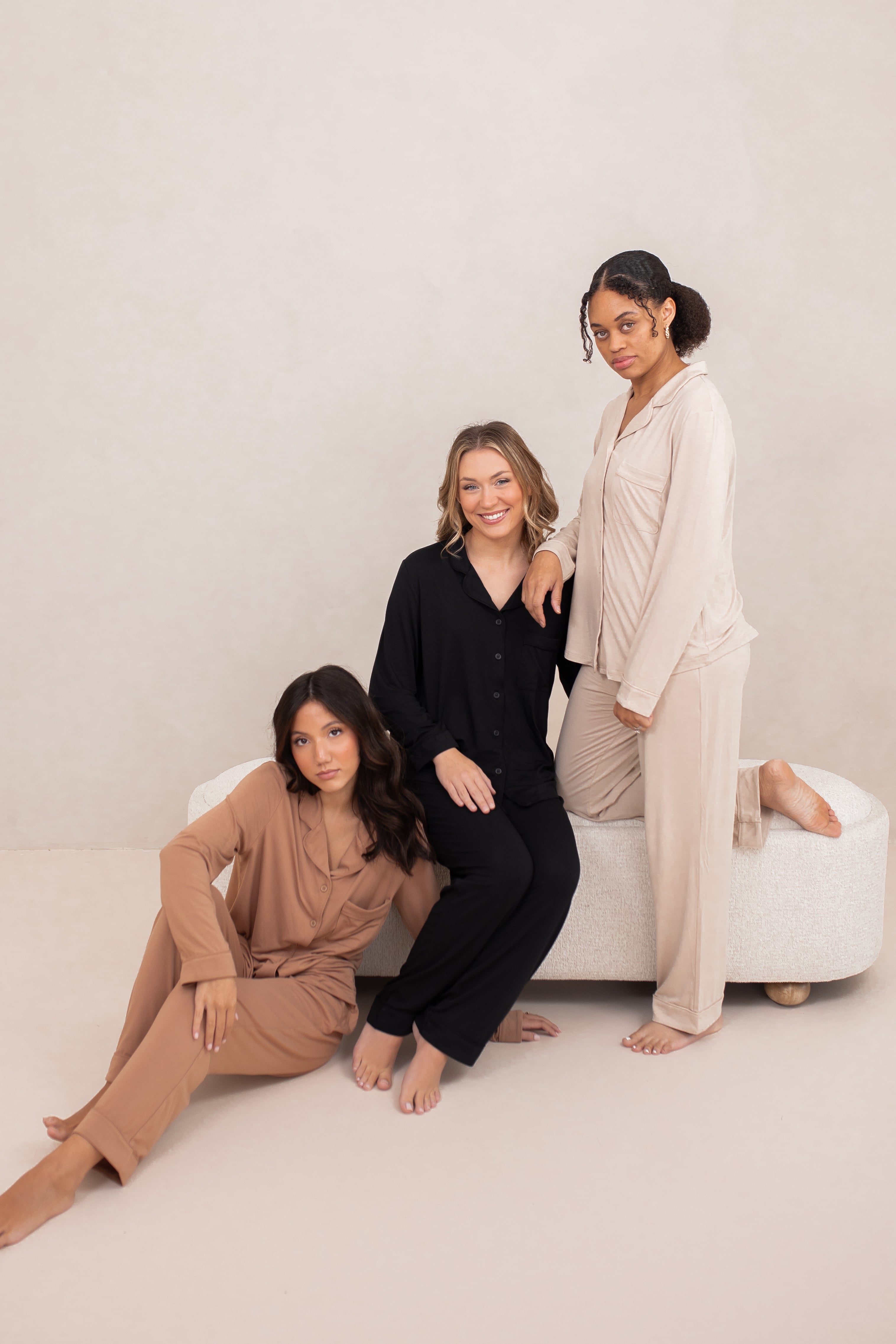 Models sitting and standing on a white couch wearing the Bamboo Long-Sleeved Women's Pajama Sets in Latte with Latte Trim, Midnight with Midnight Trim and in Bisque with Bisque Trim
