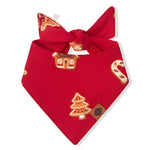 A red pet bandana featuring a festive Christmas print with gingerbread houses, candy canes, and a Christmas tree, tied in a knot at the top.