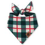 A dog bandana bib with a green, white, and red plaid pattern, featuring a tie at the top and a small brown leather patch on the bottom right corner, shown on a plain white background.