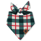 A dog bandana bib with a green, white, and red plaid pattern, featuring a tie at the top and a small brown leather patch on the bottom right corner, shown on a plain white background.