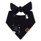 A black dog bandana with a bow, decorated with small Christmas-themed illustrations including a pine tree, snowman, stars, and a gold spoon, shown on a plain white background.