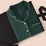 A folded dark green pajama shirt with white piping, presented in a black gift box on a light beige background.