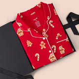 A red long-sleeve pajama shirt with white piping and a holiday print of gingerbread houses, trees, and candy canes, presented in an open black gift box on a beige background.