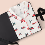 A folded white pajama shirt with red piping and a repeating print of red pickup trucks and dark green Christmas trees, displayed on a black gift box with a black ribbon against a pale pink background.
