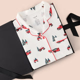 A folded white pajama shirt with red piping and a repeating print of red pickup trucks and dark green Christmas trees, displayed on a black gift box with a black ribbon against a pale pink background.
