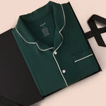 A dark green pajama shirt with white piping trim, folded and presented in a black gift box with a black ribbon on a light beige background.