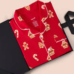 A red long-sleeve pajama shirt with white piping and gingerbread-themed prints, presented in an open black gift box on a light beige background.