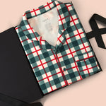 A folded long-sleeve plaid pajama shirt in green, white, and red, presented in a black gift box with a black ribbon on a solid pale pink background.