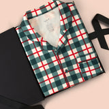 A folded long-sleeve plaid pajama shirt in green, white, and red, presented in a black gift box with a black ribbon on a solid pale pink background.