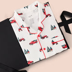 A folded white pajama shirt with red piping and a red Christmas tree tractor print, presented on a black gift box with a black ribbon against a light pink background.