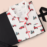 A folded white pajama shirt with red piping and a red Christmas tree tractor print, presented on a black gift box with a black ribbon against a light pink background.