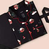 A folded black pajama set with a Christmas village print, featuring red piping and a button-down front, displayed on a black gift box with a ribbon against a light beige background.