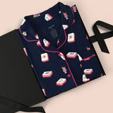 A navy blue pajama set with pink piping and a playful print of red, white, and blue mahjong tiles, presented in an open black gift box on a light pink background.