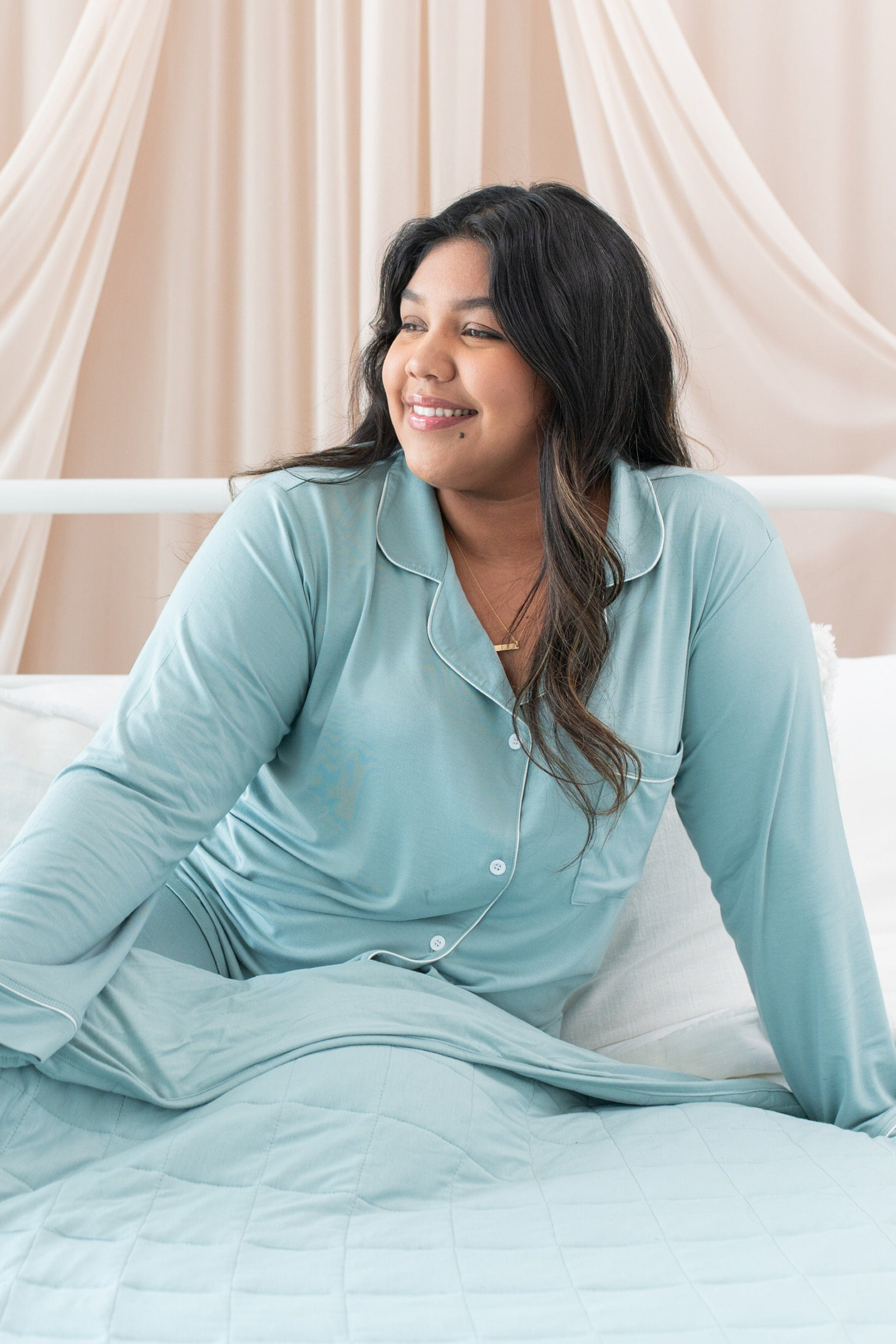 Woman wearing a light blue satin pajama set sitting on a bed with a neutral background