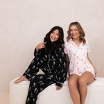 Smiling models sitting on a white bench while wearing the Bamboo Women's Short Sleeve Pajama Set in Pastel Ghost and Bamboo Women's Short Sleeve Pajama Set in Skeleton