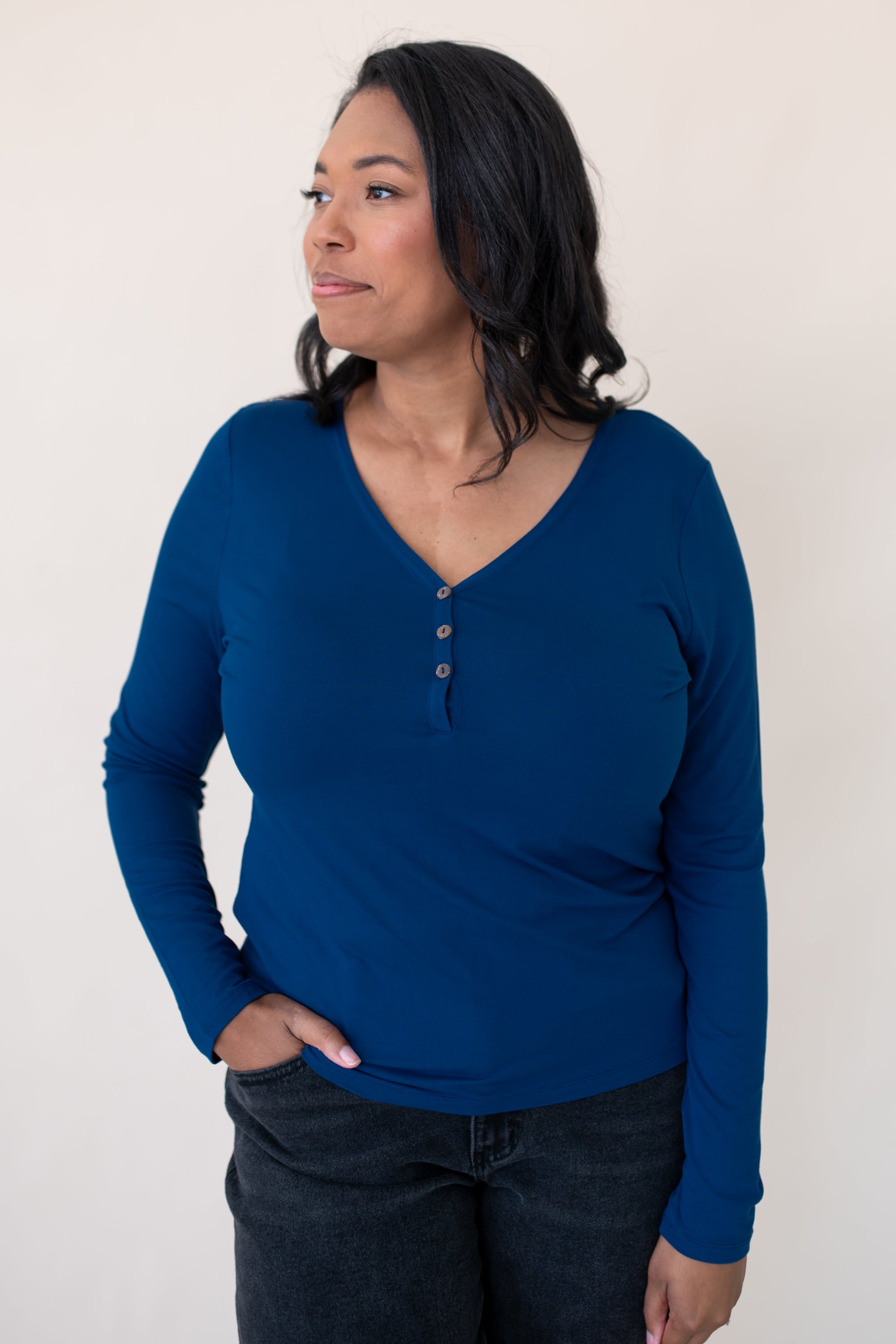 Model looking to the side while wearing the Women's Long Sleeve Henley Tee in Tahoe paired with dark denim jeans