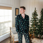 Man standing in a white paneled room with christmas trees while modeling the Kyte Men's Long-Sleeve Pajama Set in Twinkle Tree