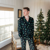 Man standing in a white paneled room with christmas trees while modeling the Kyte Men's Long-Sleeve Pajama Set in Twinkle Tree