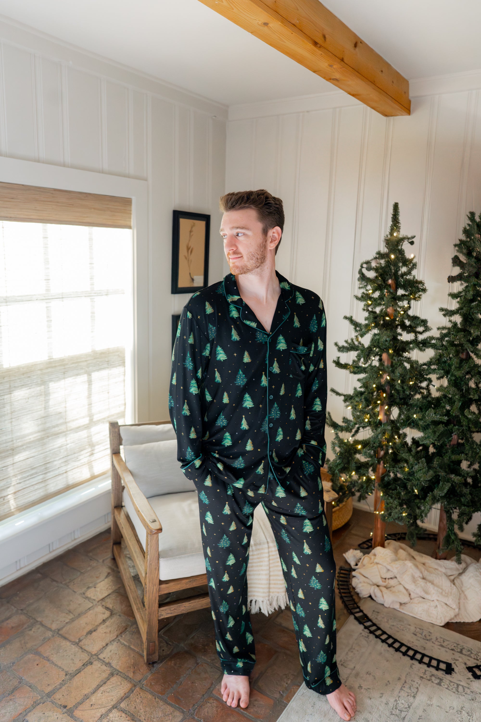 Man standing in a white paneled room with christmas trees while modeling the Kyte Men's Long-Sleeve Pajama Set in Twinkle Tree