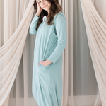 Woman wearing a light blue sleep bag walker standing in front of sheer curtains.