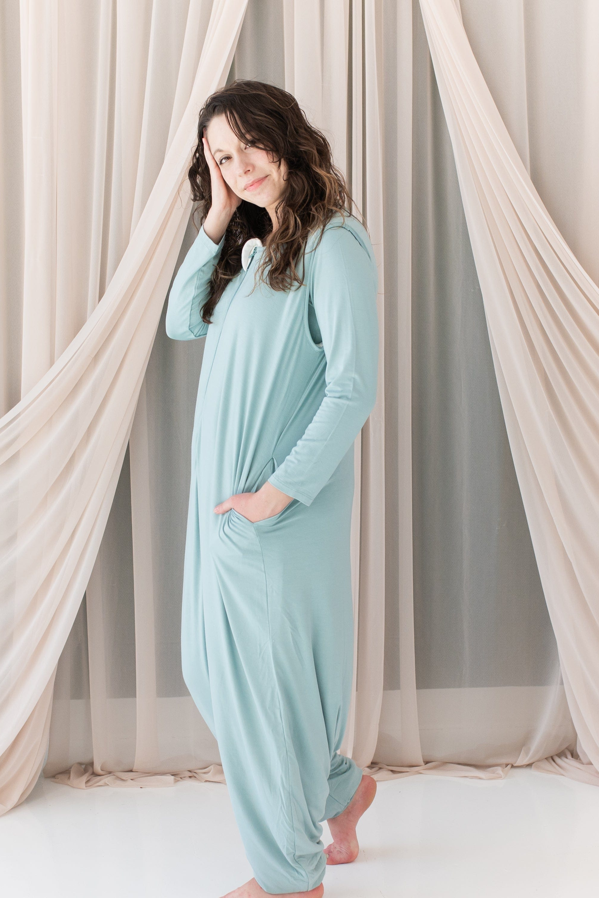 Woman wearing a light blue sleep bag walker standing in front of sheer curtains.