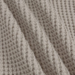Close-up of a textured knit fabric with a waffle or basketweave pattern in a warm taupe-beige color, shown with soft folds.