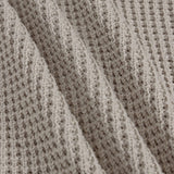 Close-up of a textured knit fabric with a waffle or basketweave pattern in a warm taupe-beige color, shown with soft folds.