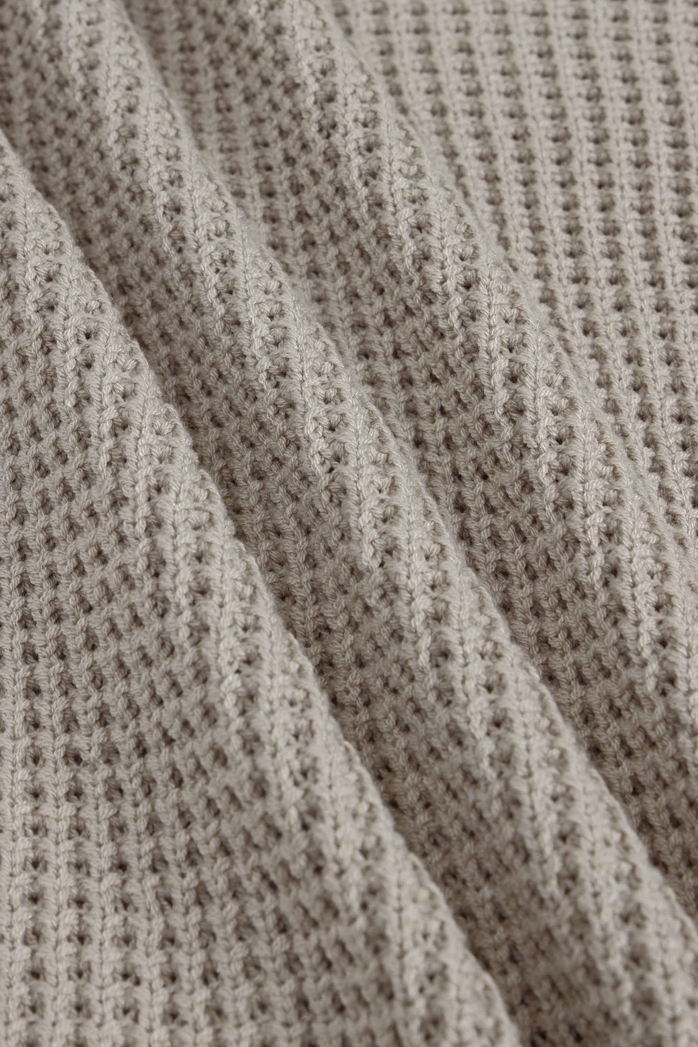 Close-up of a textured knit fabric with a waffle or basketweave pattern in a warm taupe-beige color, shown with soft folds.