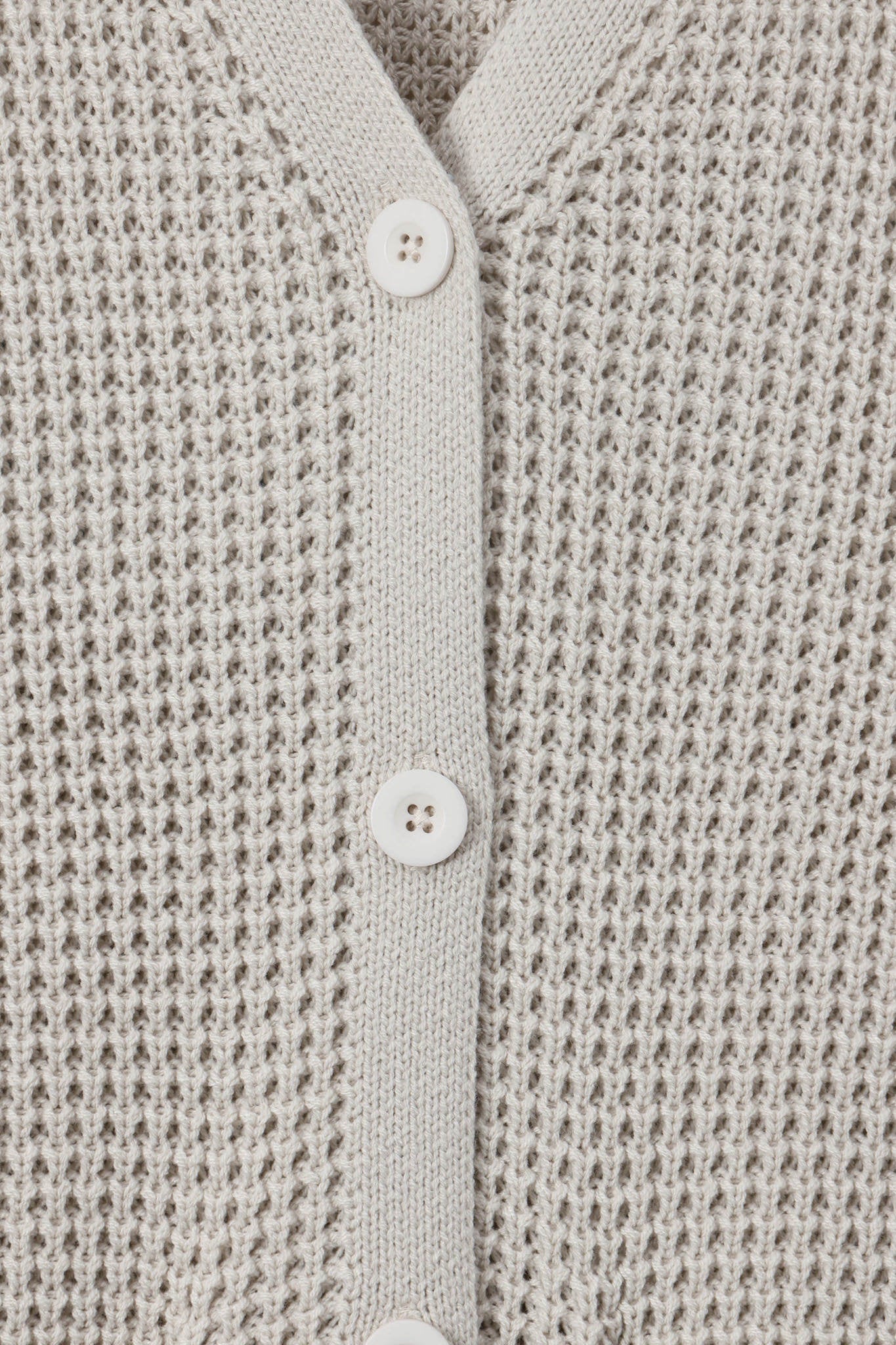 Close-up of a textured knit cardigan in a light warm gray or oatmeal tone, featuring a button placket with white buttons.