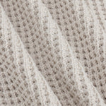 Close-up of a textured knit fabric in a warm off-white or cream tone, showing the detailed weave and soft folds.