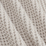 Close-up of a textured knit fabric in a warm off-white or cream tone, showing the detailed weave and soft folds.