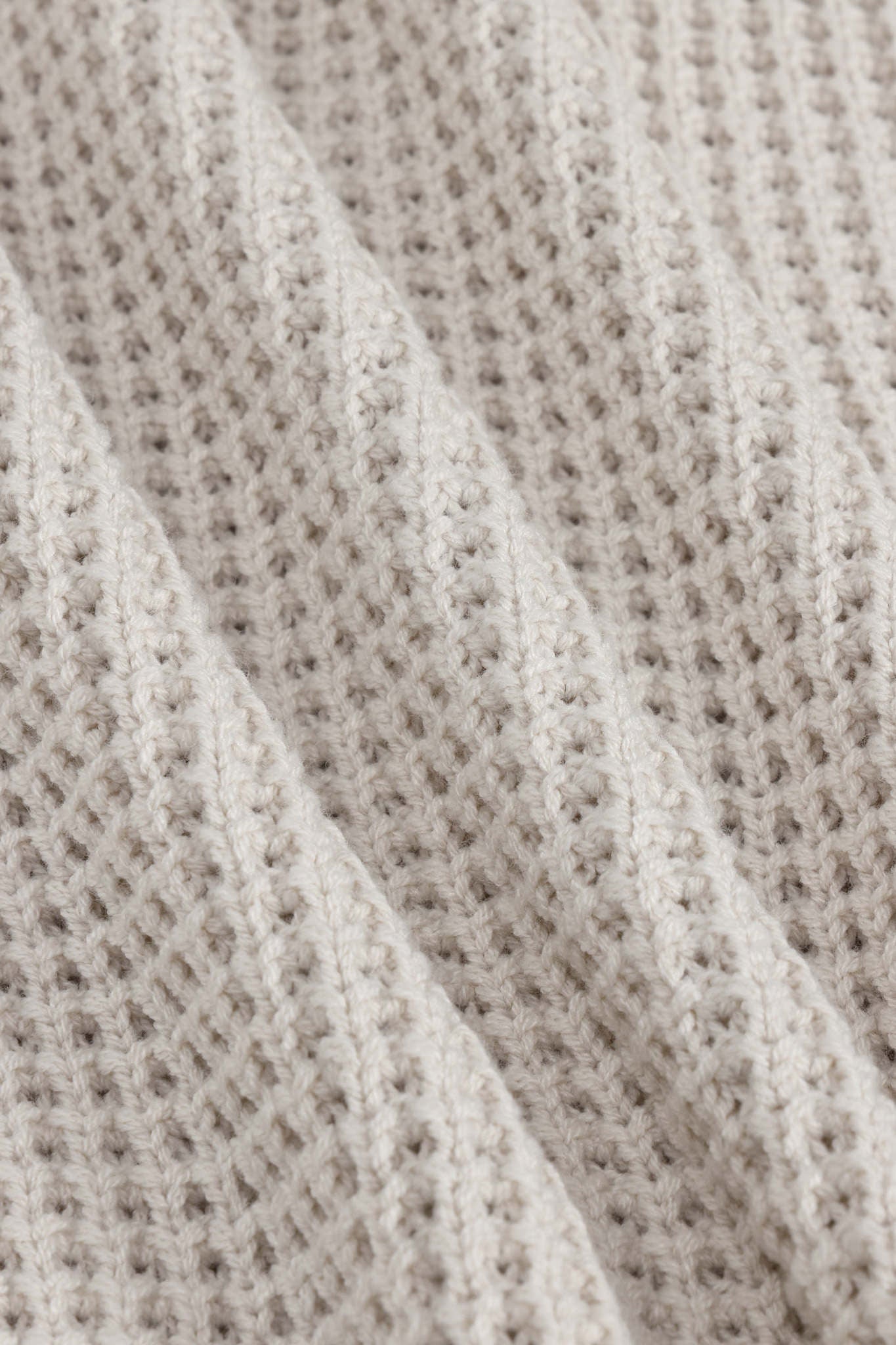 Close-up of a textured knit fabric in a warm off-white or cream tone, showing the detailed weave and soft folds.