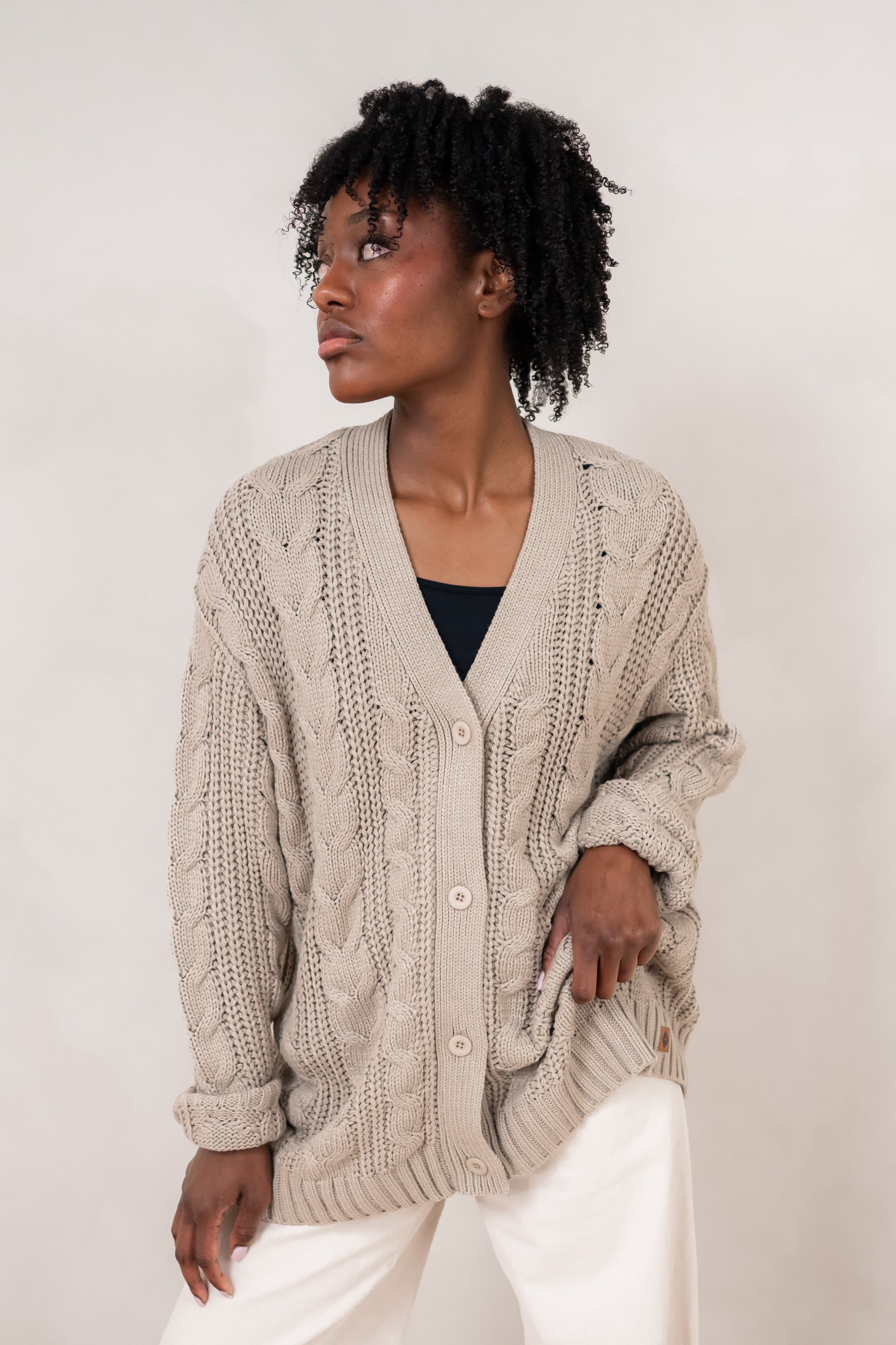Model looking to the side while wearing the Chunky Knit Cable Cardigan in Almond paired with white pants