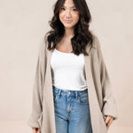Woman wearing a tan cardigan, white top, and blue jeans against a plain background
