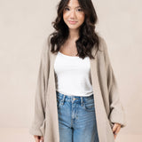 Woman wearing a tan cardigan, white top, and blue jeans against a plain background